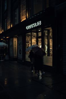 Moody nighttime street scene with people under umbrellas in front of a glowing storefront.