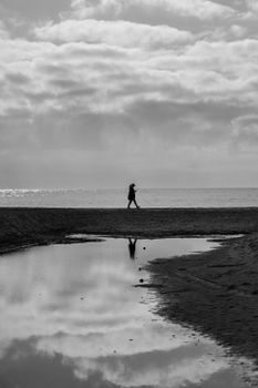 Monochrome image of a woman walking on a seashore in Palafrugell, Spain, under a cloudy sky.