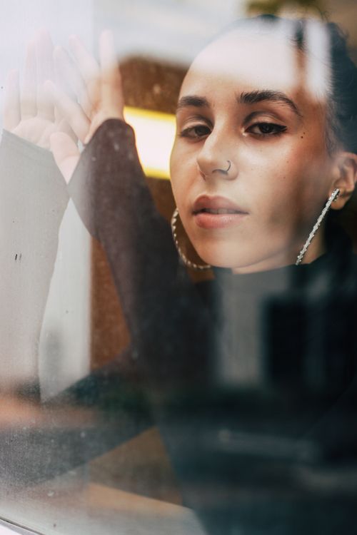 Free Fashionable young woman in a black top posing behind a glass window, reflecting urban elegance. Stock Photo