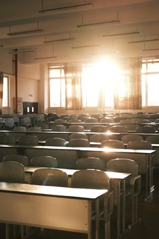 A warmly lit empty classroom with sunlight streaming through windows, illuminating rows of desks.