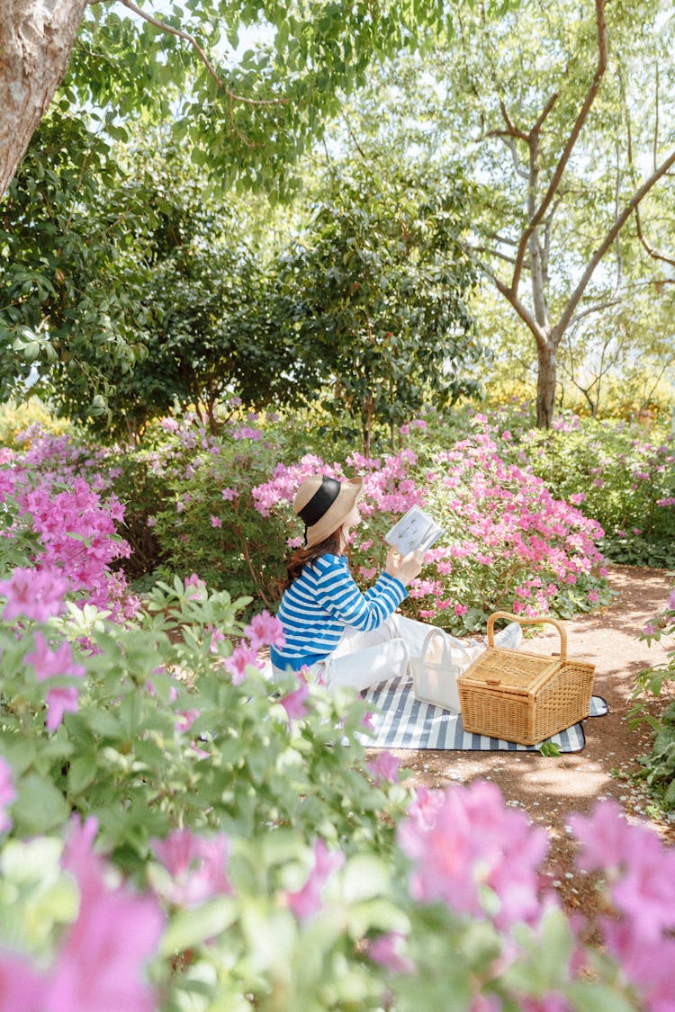 Woman Sitting On A Blanket In A Park Among Flowers And Reading A Book 