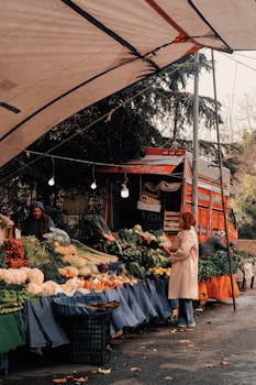 A bustling outdoor vegetable market with shoppers examining fresh produce under a canopy.