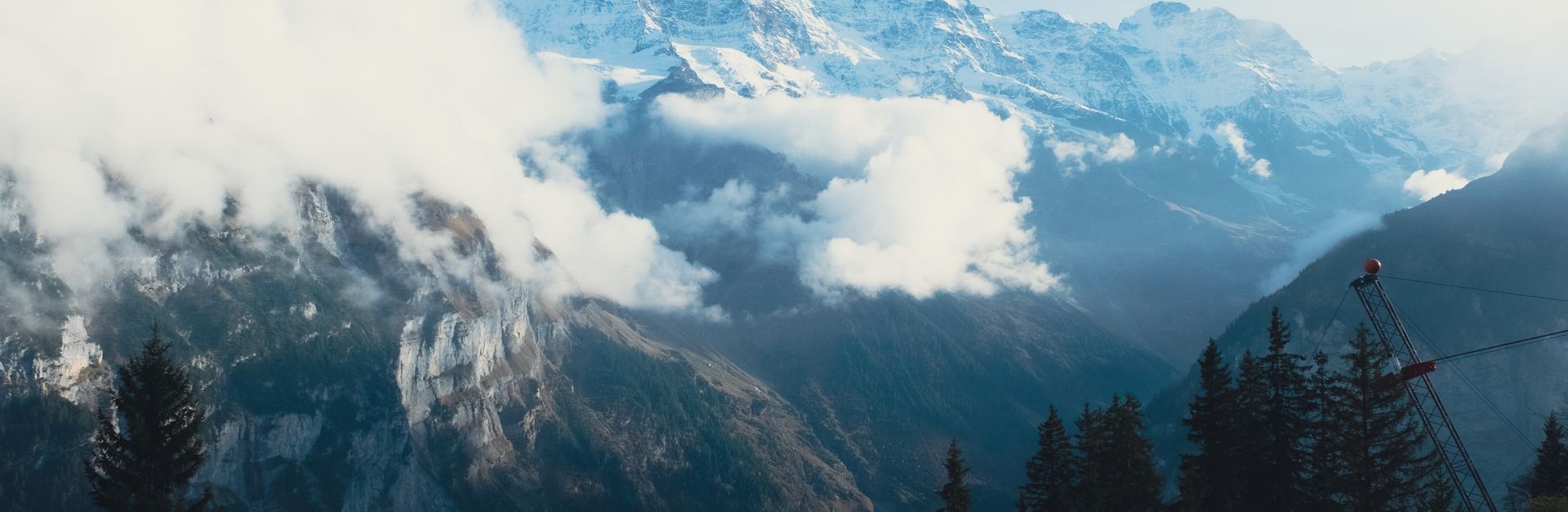 A breathtaking view of snowy mountain peaks partially shrouded in clouds with pine trees in the fore