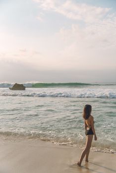 Woman in black bikini stands on a serene beach, gazing at the ocean waves.