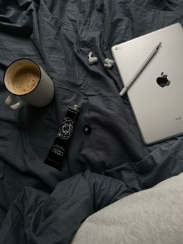 Flat lay of coffee, tablet, earphones, and face cream on a bed, creating a cozy morning vibe.