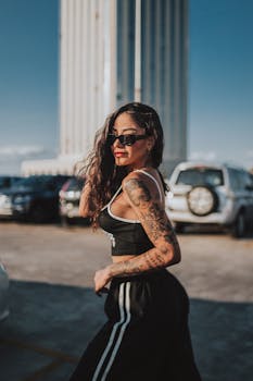 Confident woman with sunglasses and tattoos strikes a pose in an urban parking lot under clear skies.