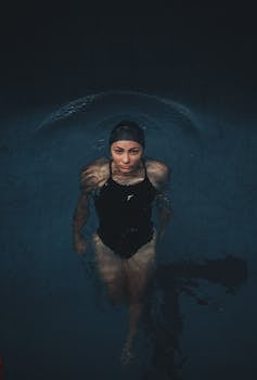 Young woman swimming with concentration in dimly lit pool wearing a black bathing suit and cap.