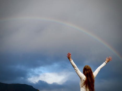 A woman with raised arms under a vibrant rainbow in Selfoss, Iceland.