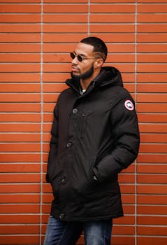 Stylish adult man wearing sunglasses and winter coat, leaning against a brick wall.