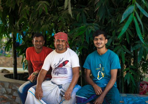 Free Three men sitting together, smiling, in an outdoor setting with bamboo plants in Dhaka. Stock Photo