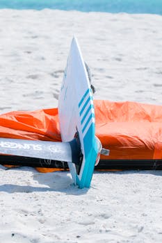 Close-up of a surfboard standing in the sand next to bright orange kite gear.