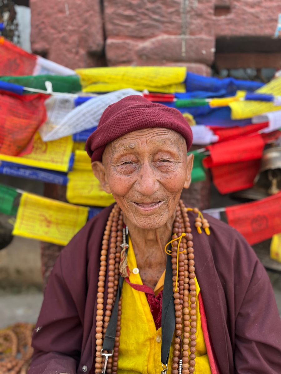Senhor sorridente em Kathmandu, com colares de contas, diante de bandeiras de oração