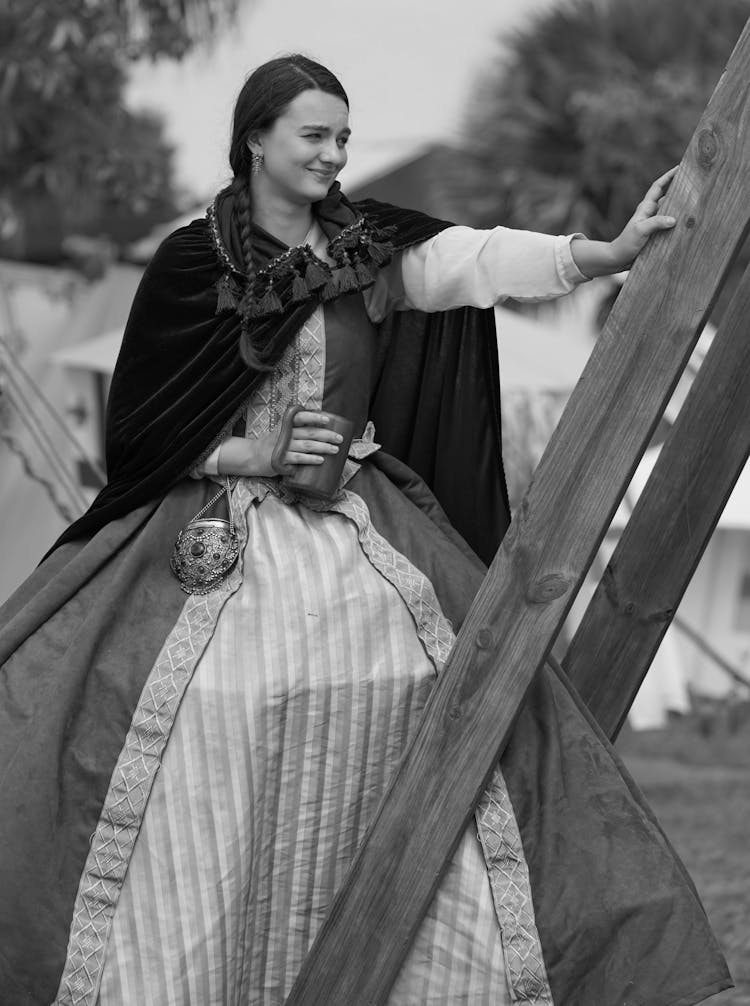 A Grayscale Photo Of A Woman In Long Dress With Cape Standing Near The Wooden Posts