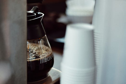 A close-up view of a coffee pot next to a stack of disposable cups in an indoor setting.