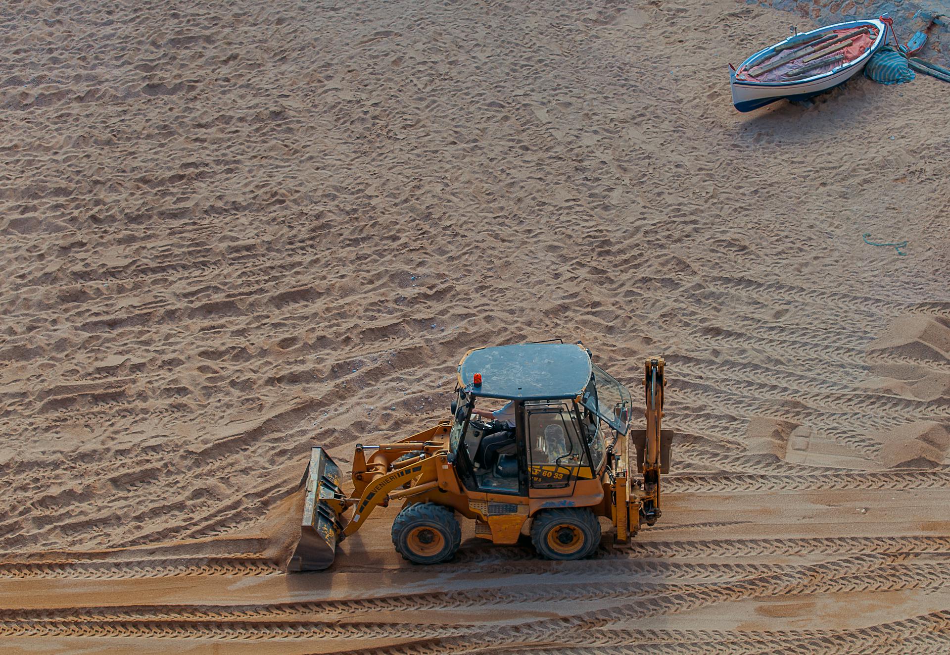 An aerial view of a backhoe loader operating on a sandy beach with a boat on the shore.