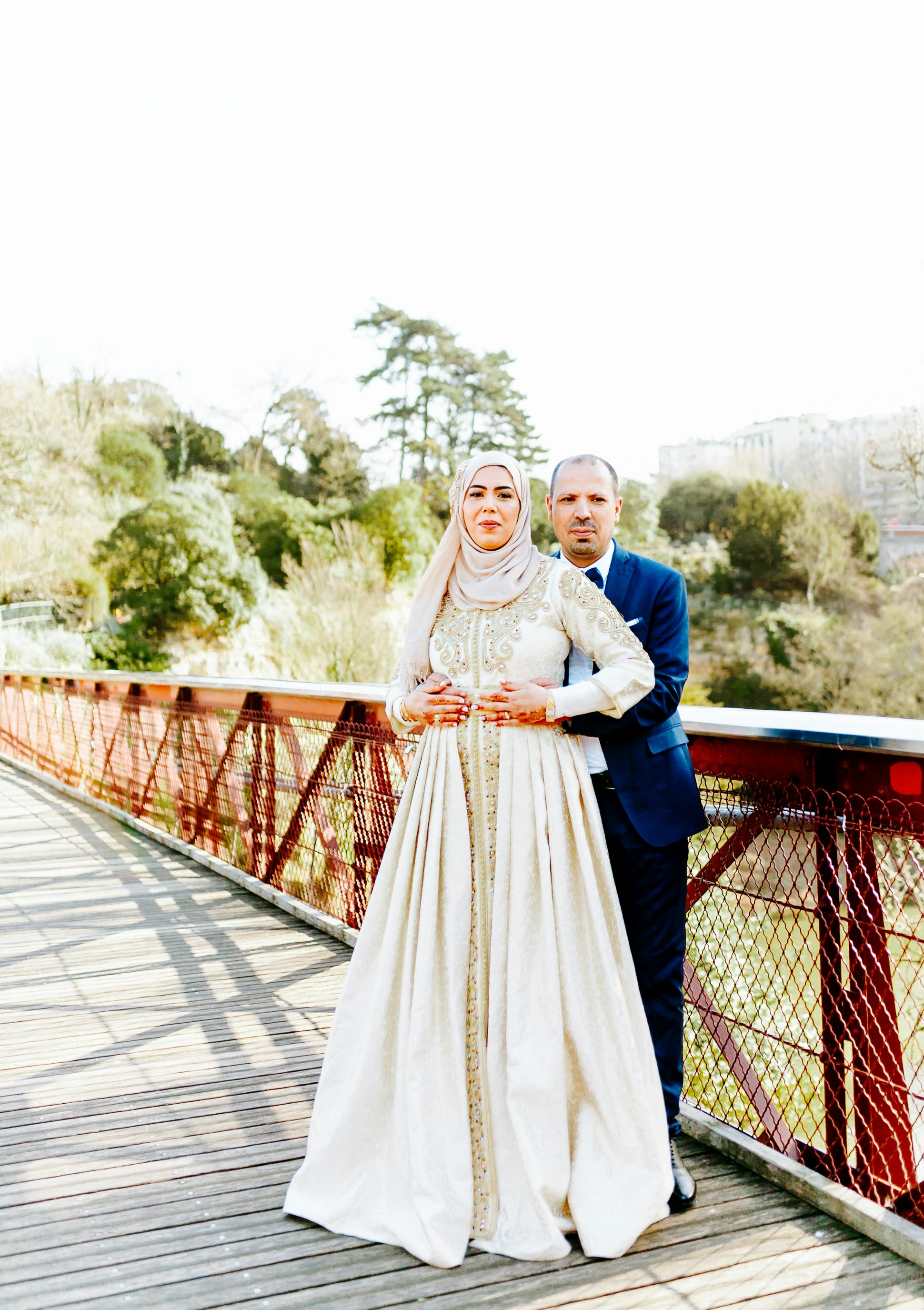 Elegant Muslim couple in wedding attire embracing on a sunny bridge in New York City, showcasing love and tradition.