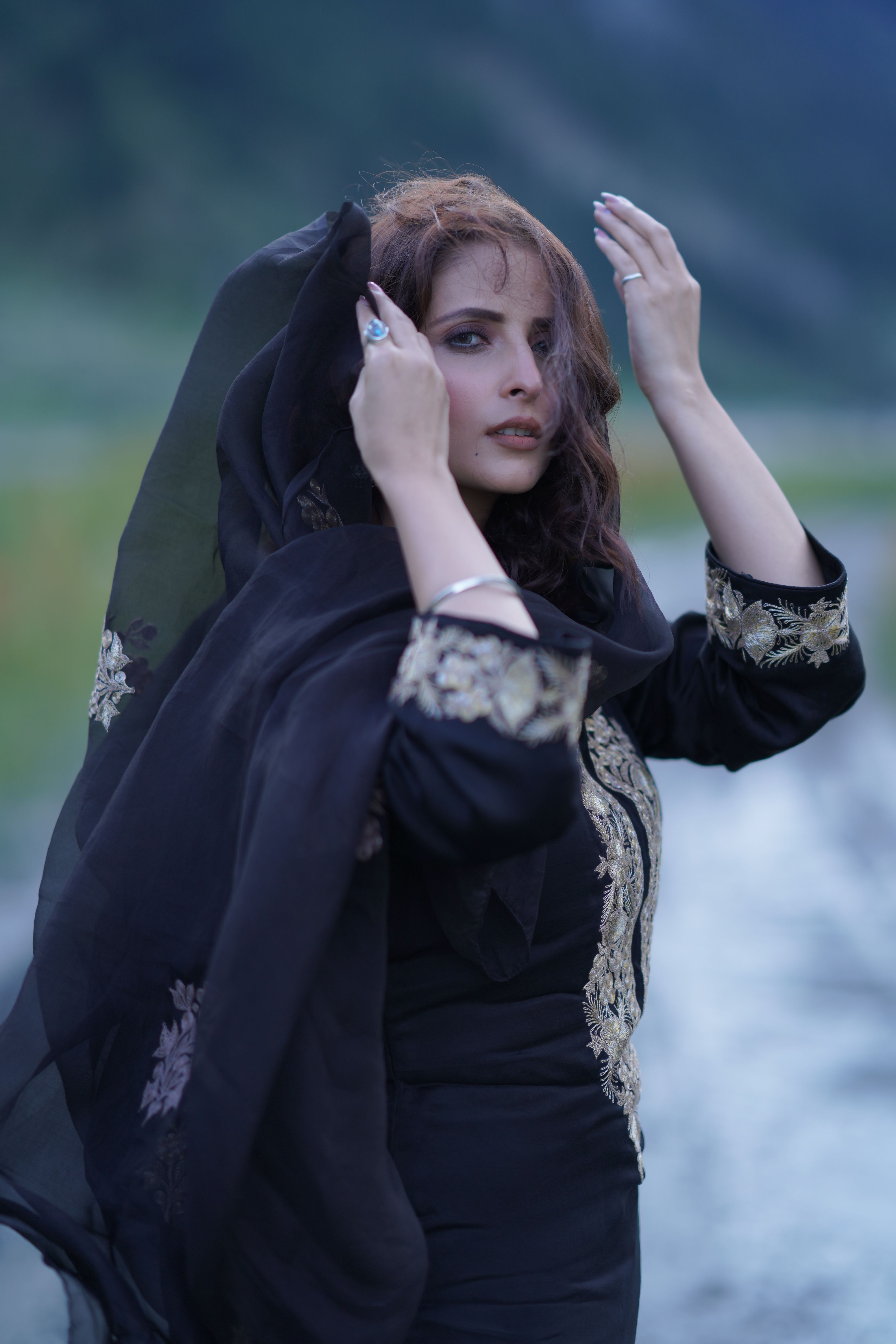 A woman wearing a black embroidered dress and headscarf poses elegantly outdoors, reflecting grace and style.