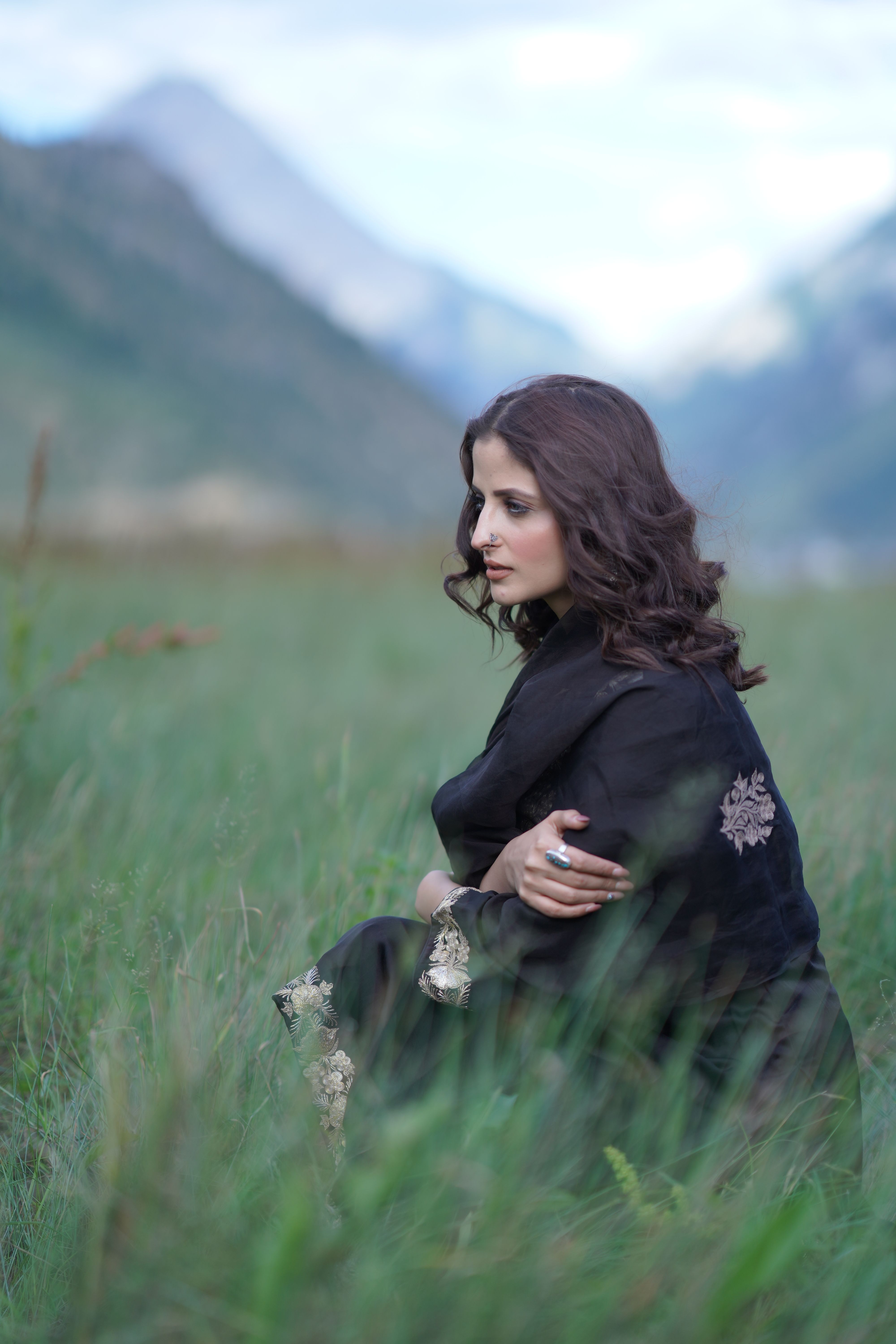 A brunette woman in elegant attire sits thoughtfully in a scenic green field.