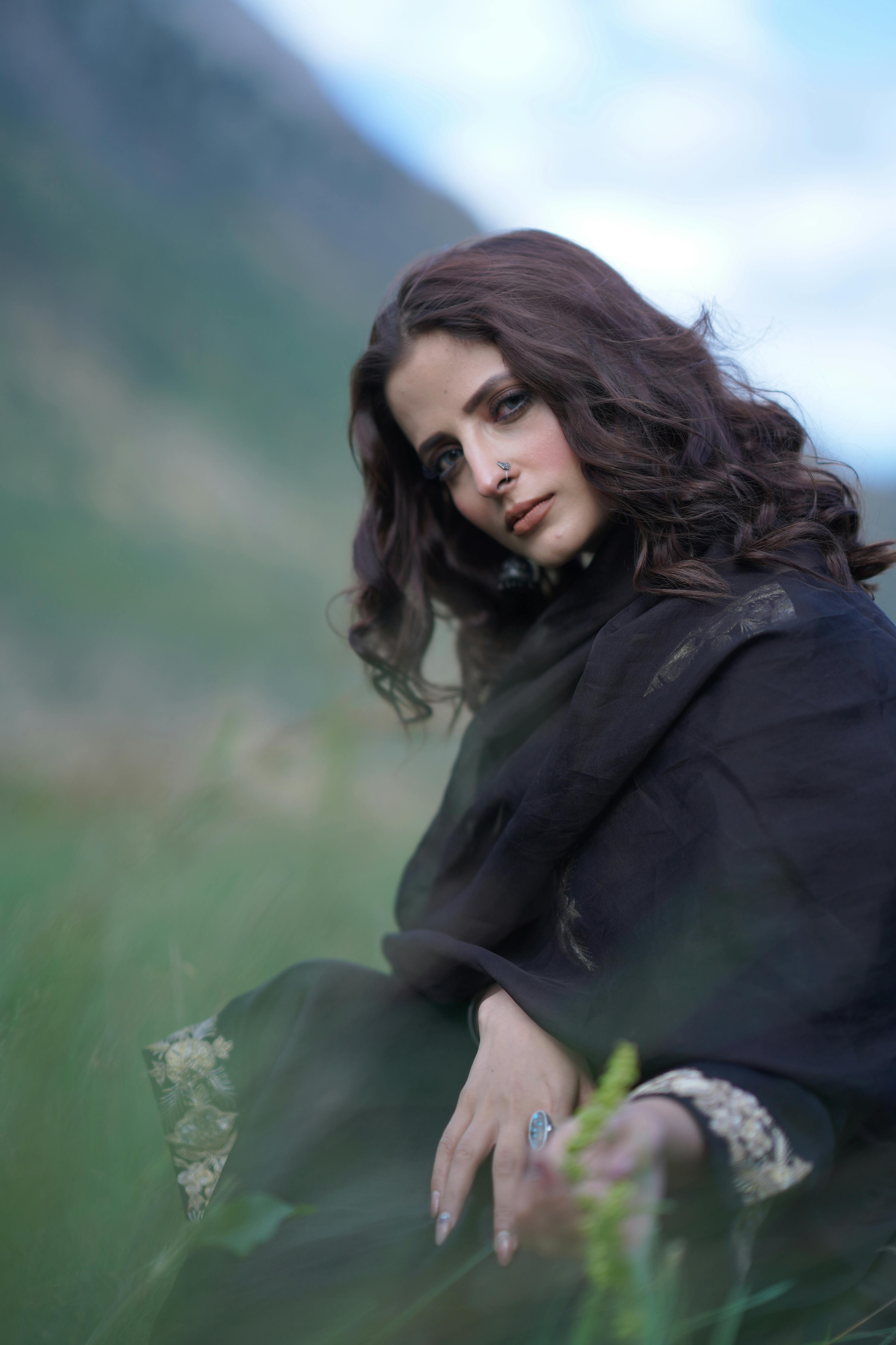 A brunette woman poses elegantly in a field, wearing a dark flowing gown. Captured in a soft, natural setting.