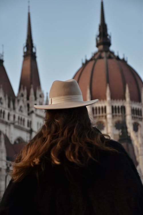 Back View Shot of a Person in Black Jacket and White Fedora Hat Standing Near Hungry Parliament Building