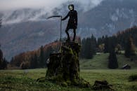 云雾缭绕的山间树桩上，站着一个戴着面具和兜帽的人，手里拿着镰刀