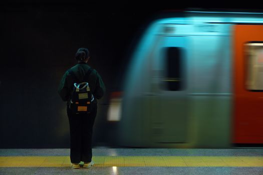 A woman with a backpack stands at an Ankara metro station as a train speeds by.