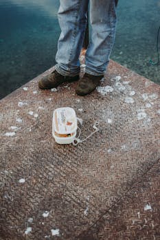 Man in jeans and boots standing next to tackle box on a metal platform by the water.