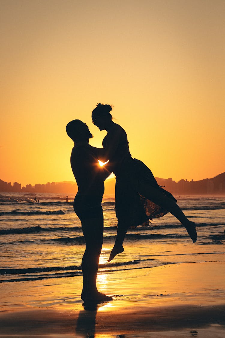 Man Picking Up A Girl On A Beach At Sunset