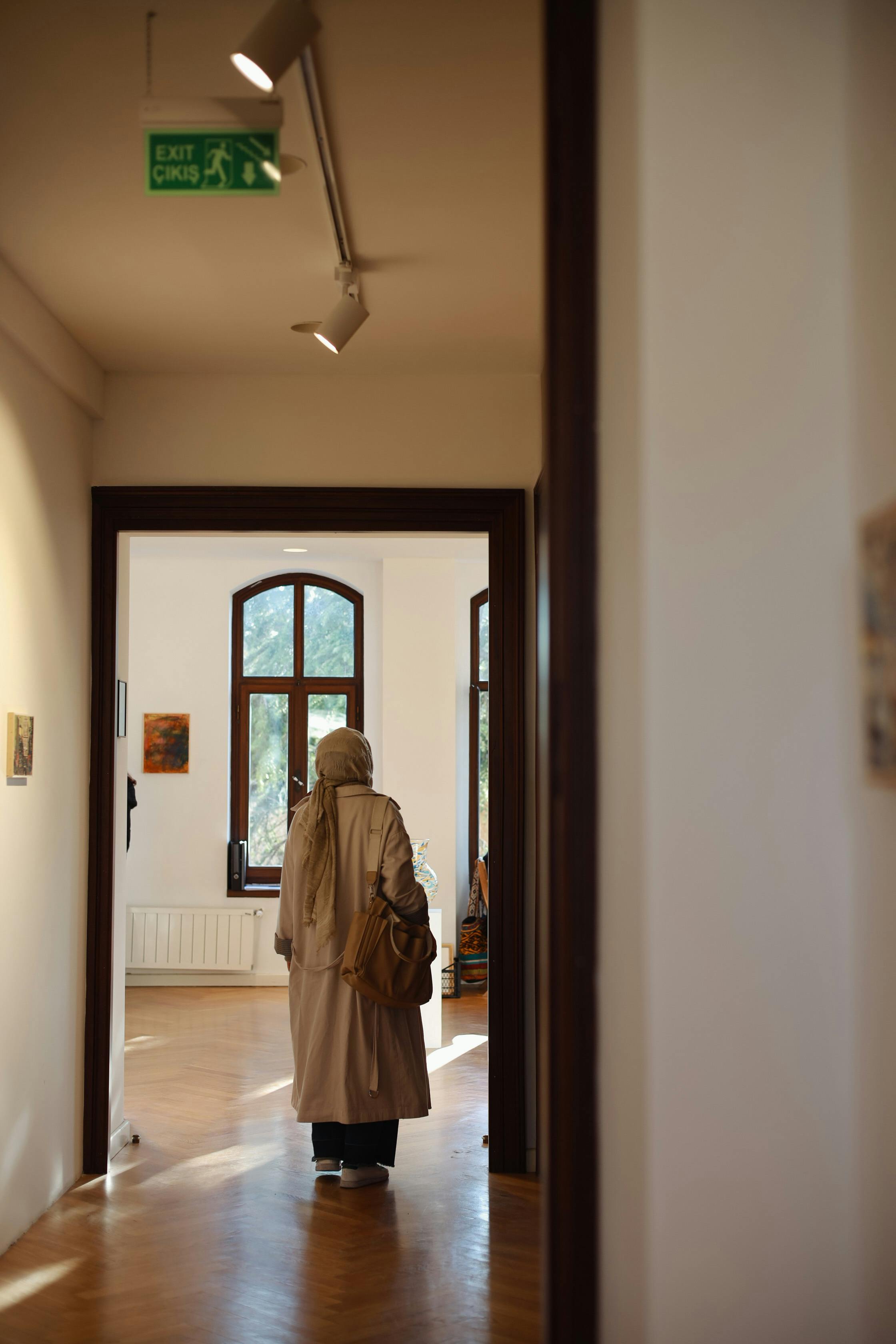 A woman in a long coat wanders through a bright art gallery, observing the paintings.