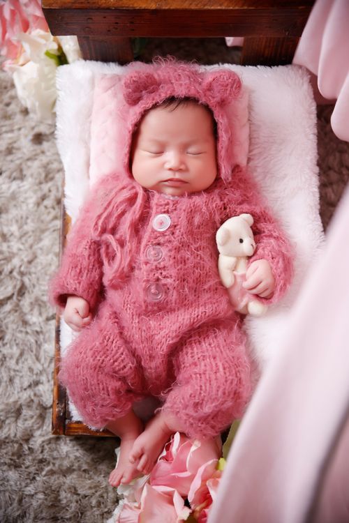 Free Charming image of a newborn baby girl asleep in a pink romper, surrounded by soft textures and a teddy bear. Stock Photo