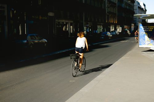 Free A woman cycles through a sunlit Paris street, showcasing urban life and transportation. Stock Photo