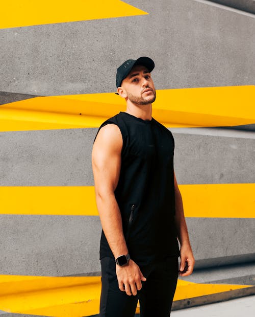 A Man in Black Tank Top and Cap Standing Near the Concrete Wall