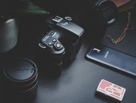 A close-up of modern photography equipment featuring a camera, lens, smartphone, and accessories on a dark surface.