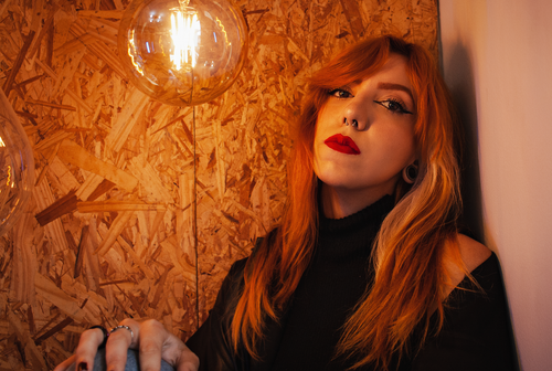 Free A close-up portrait of a redhead woman with striking red lips and piercings, leaning against an artistic wall. Stock Photo