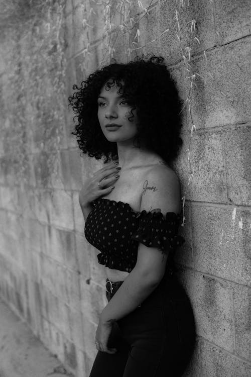 Grayscale Photo of a Woman in Black Off Shoulder Crop Top Leaning Against a Concrete Wall
