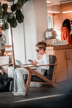 Adult woman multitasking with a laptop and phone in a stylish cafe setting.