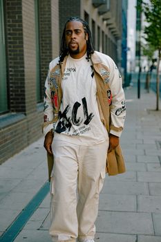 A fashionable man with braids walks down a city street in a stylish beige outfit, showcasing urban street fashion.