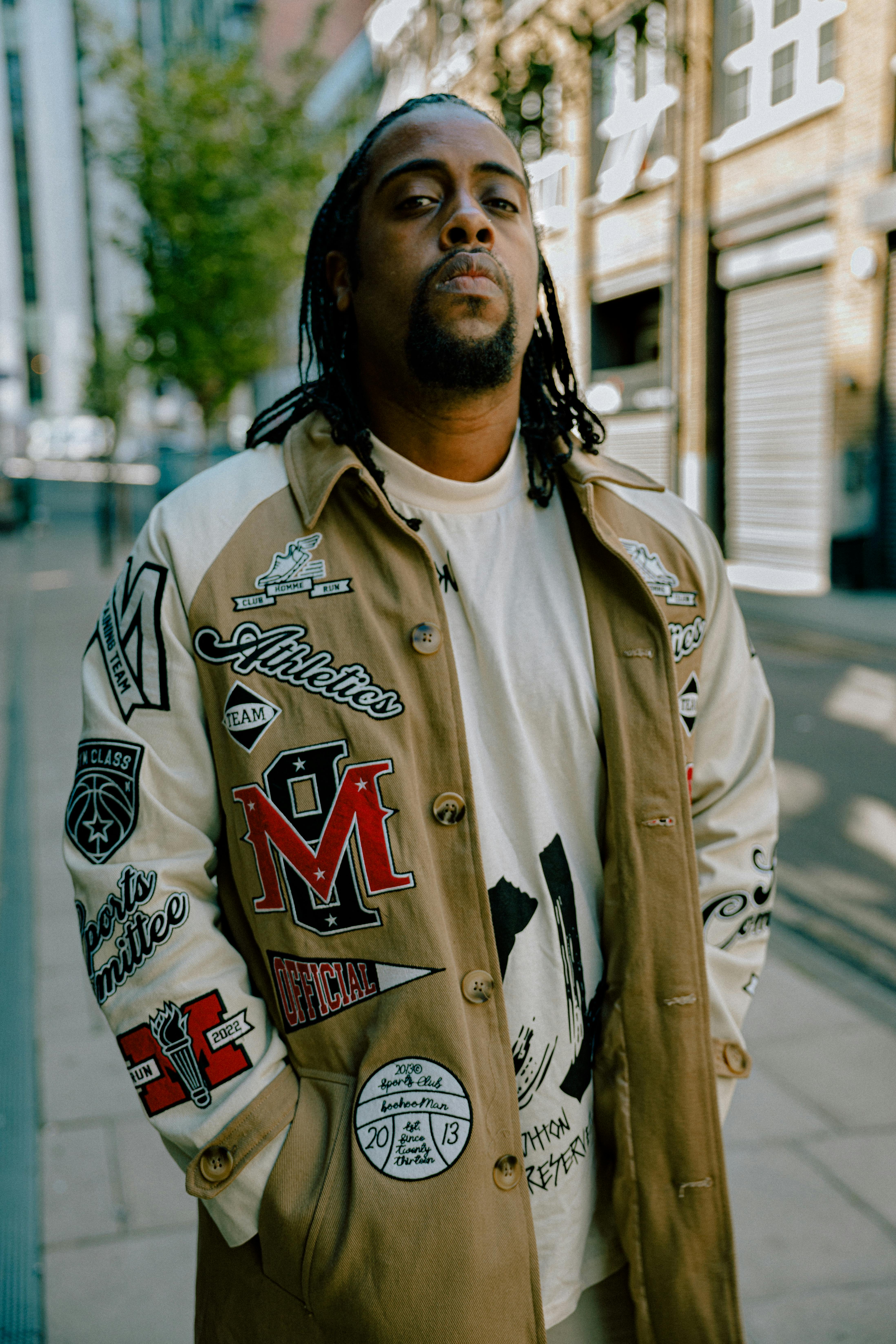 Stylish man with braids wearing a printed jacket poses confidently in an urban setting.