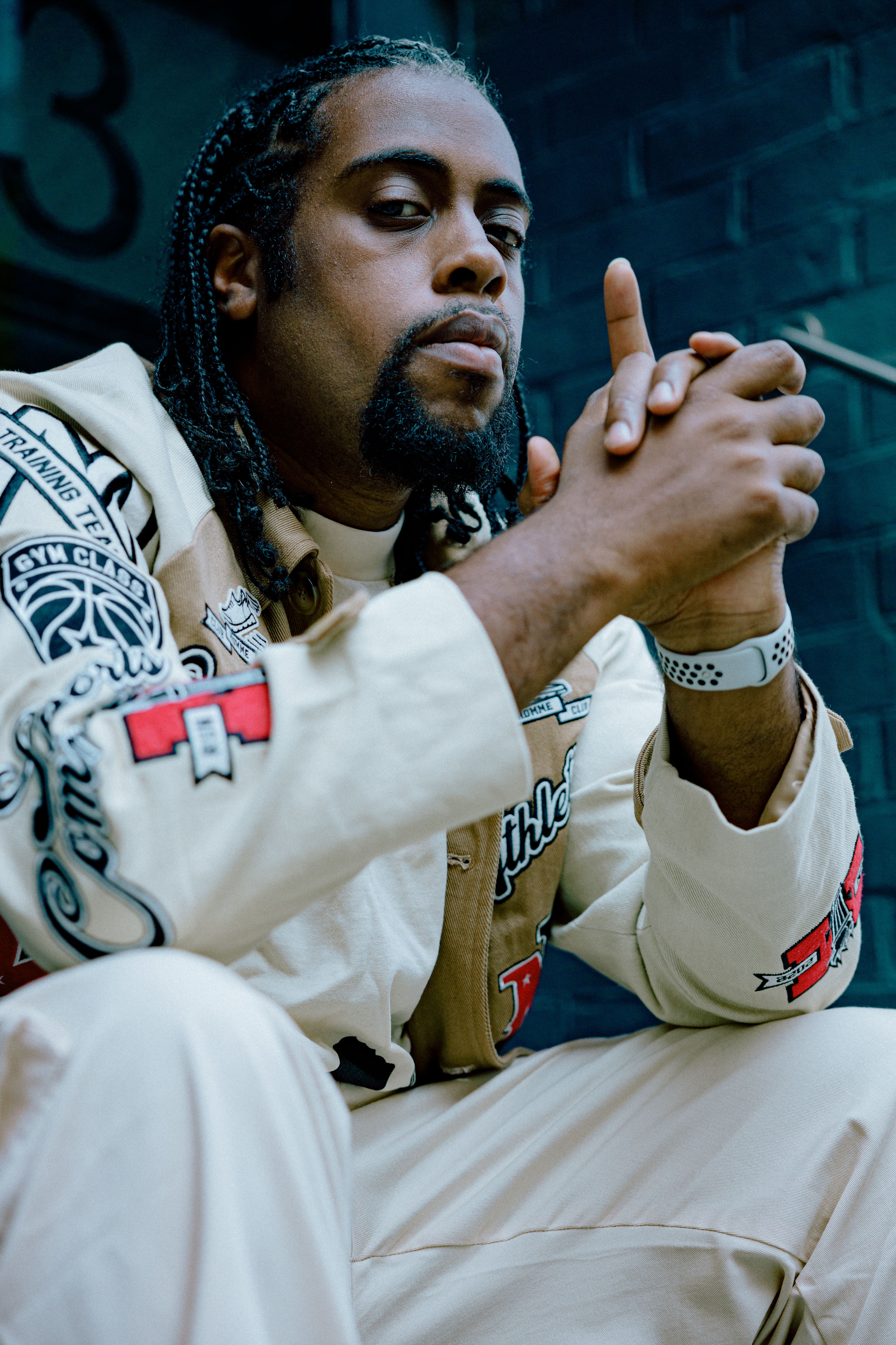 A man with braids and a serious expression sits outdoors in a fashionable embroidered outfit.