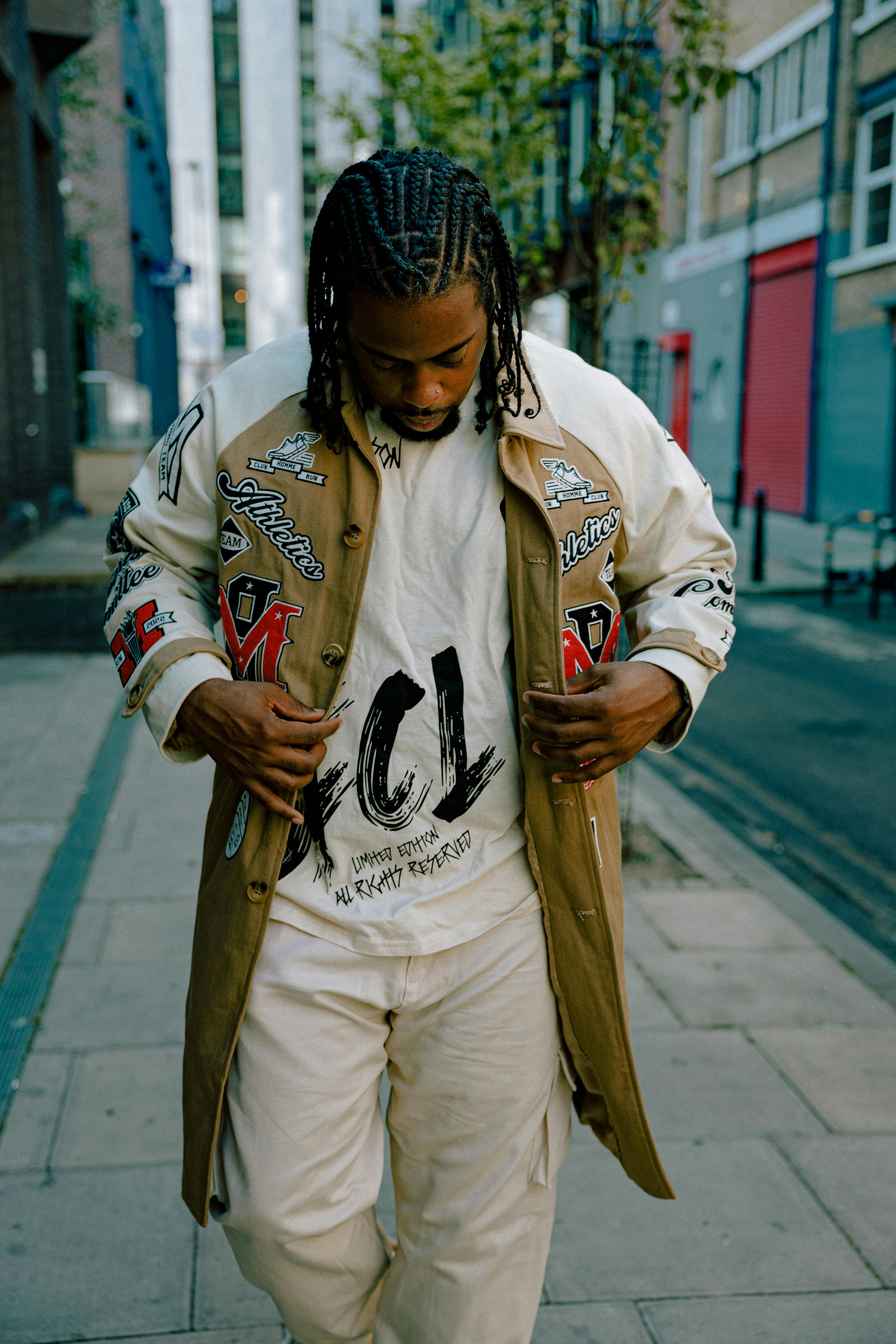 Stylish man with braids and printed jacket walking down a city sidewalk.
