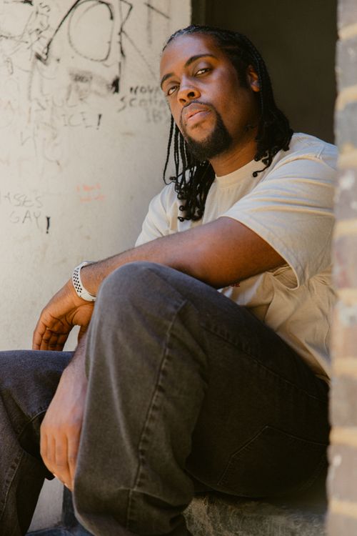 Free Portrait of a man with braided hair and a beard sitting against a graffiti wall. Stock Photo