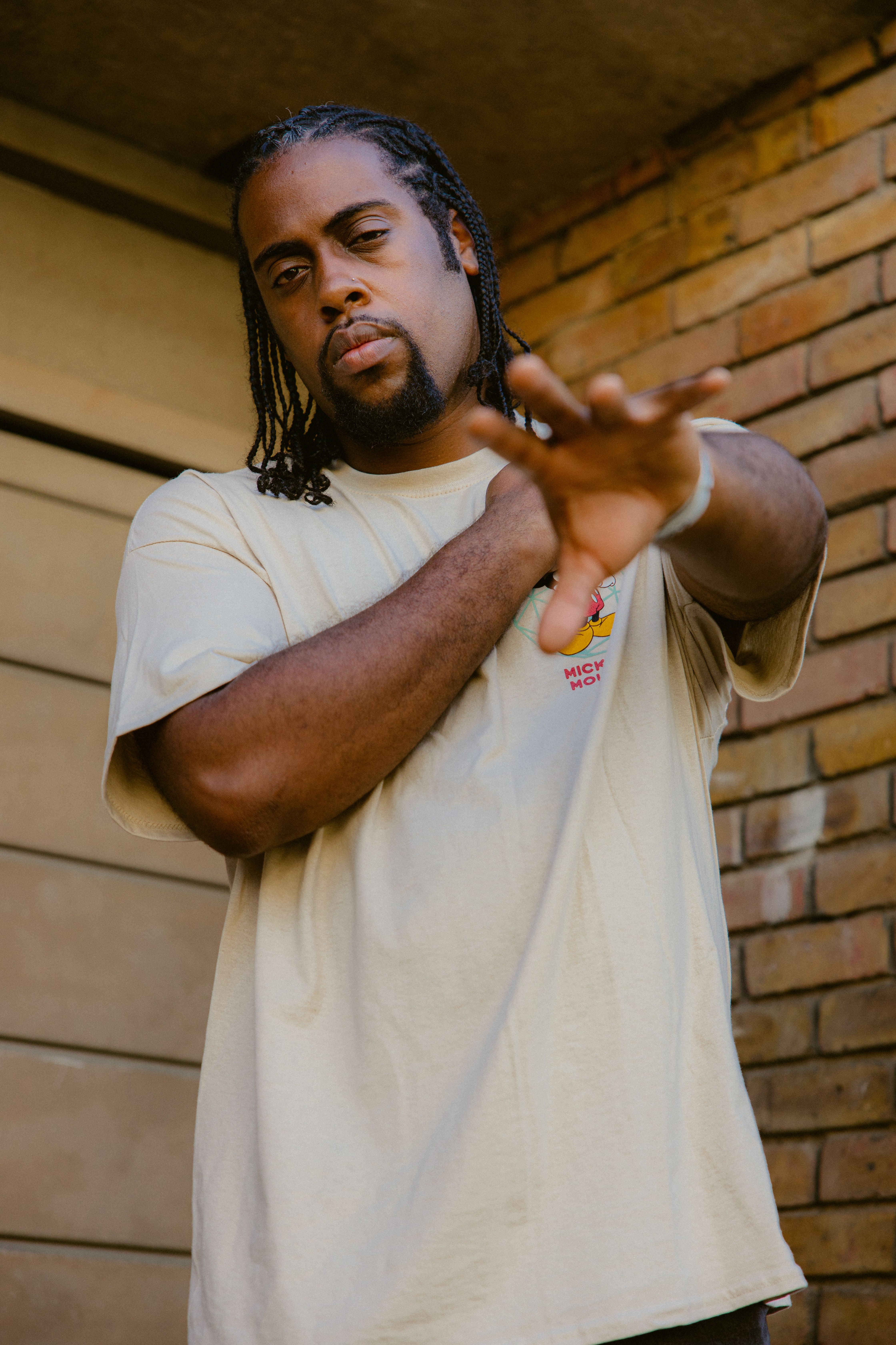 A man with braids in a white shirt posing outdoors against a brick wall.