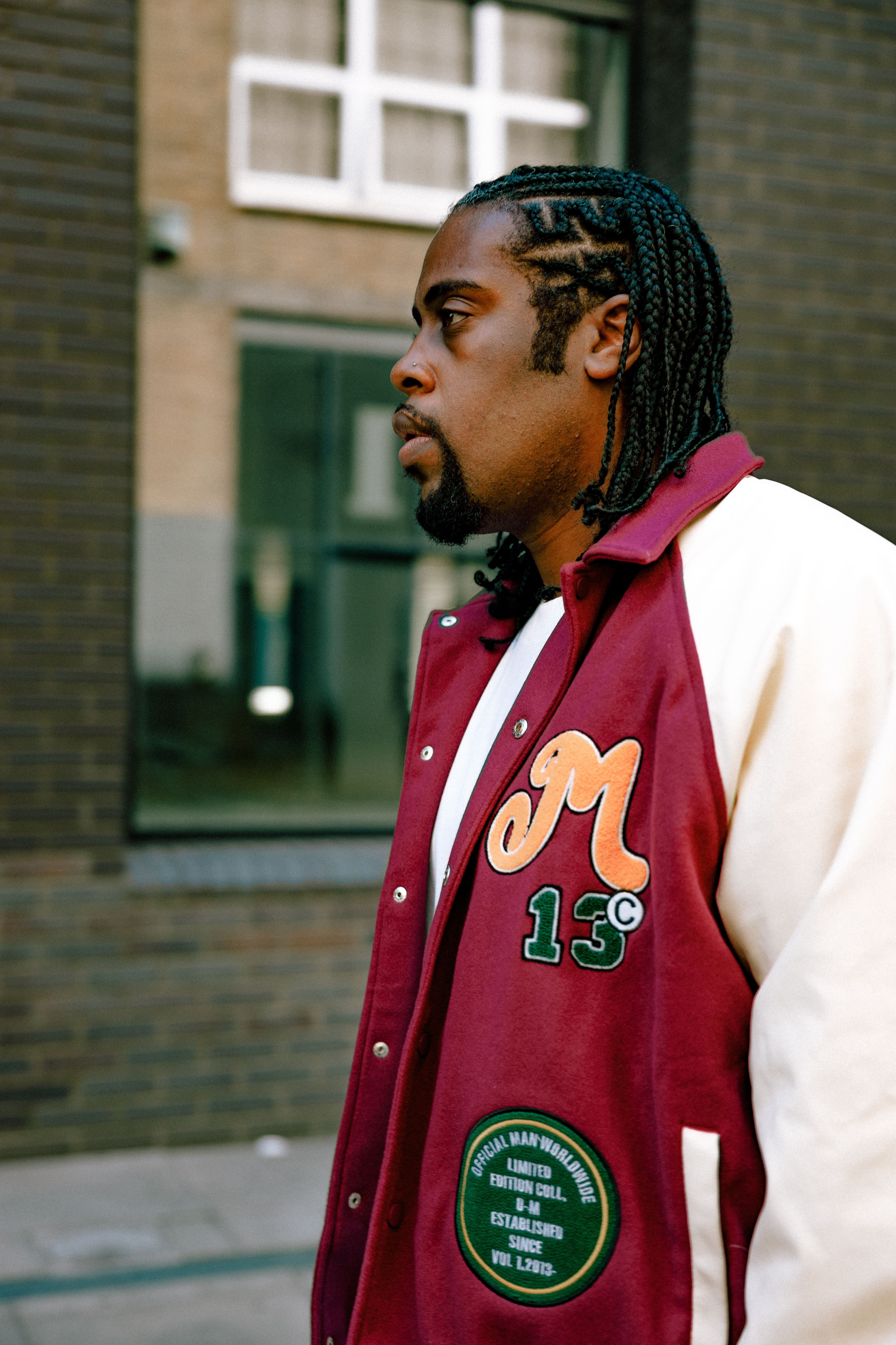 Side view of a man with braided hair wearing a varsity jacket standing outdoors.