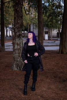 Stylish woman with purple hair stands confidently among tall trees, showcasing modern fashion.