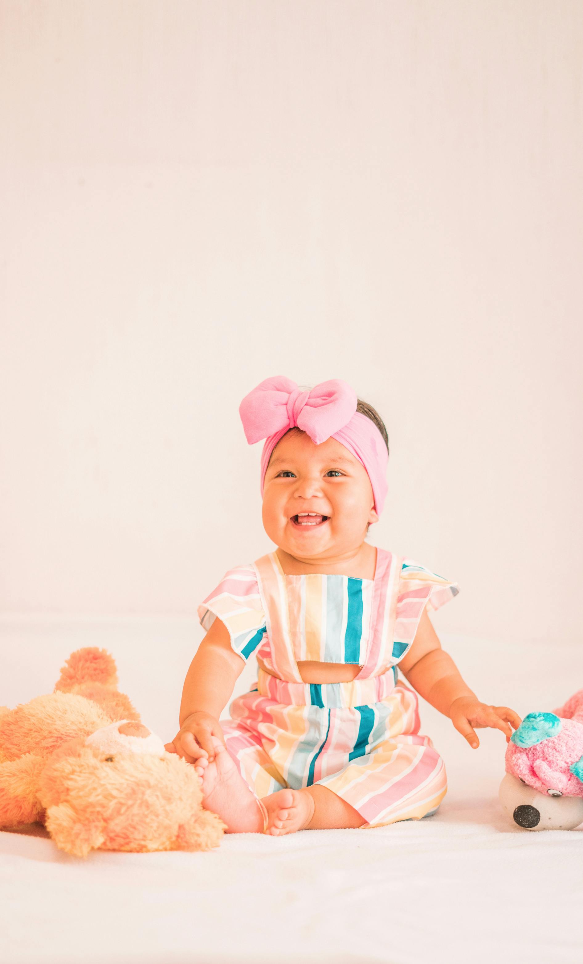 A smiling baby wearing a pink headband and colorful outfit, sitting with plush toys in a studio setting.