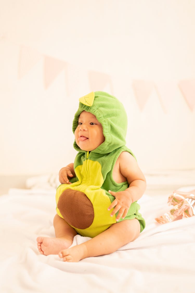 Baby In Green Avocado Costume Sitting On Bed With Tongue Out