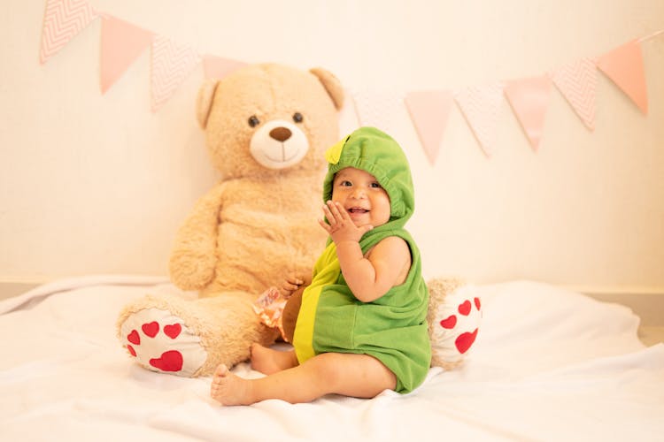 Baby Wearing Green Hoodie Sitting Next To Teddy Bear