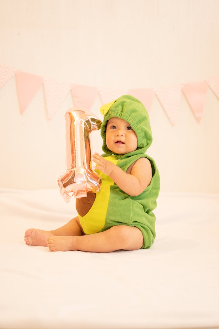 Baby In Green Avocado Romper Holding An Inflatable Balloon