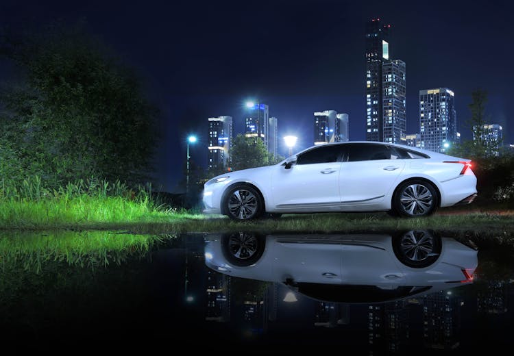 A White Car Parked On Green Grass Field Near The Body Of Water And Buildings At Night