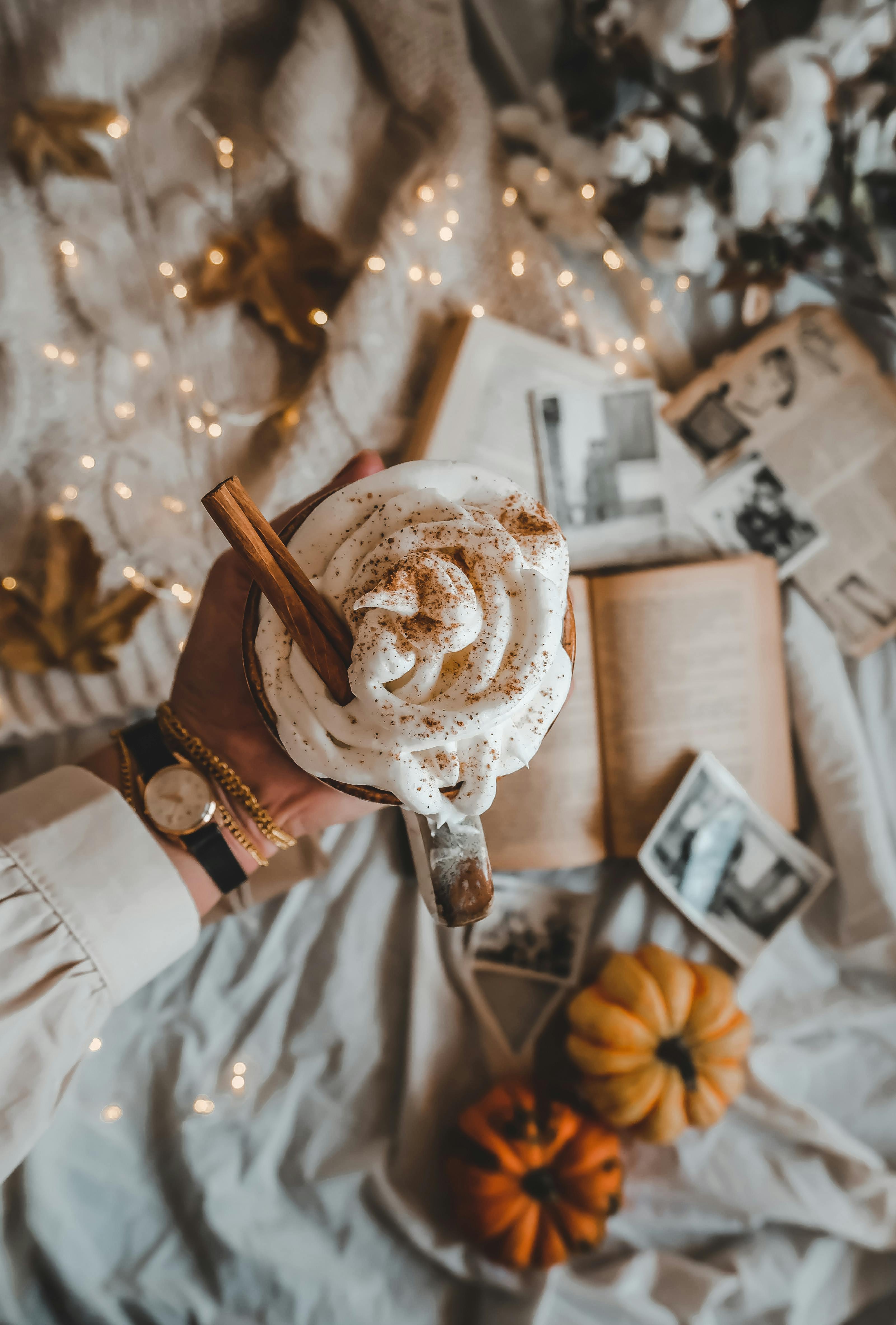 Hand holding a warm cinnamon-spiced drink with whipped cream, surrounded by autumn decor.